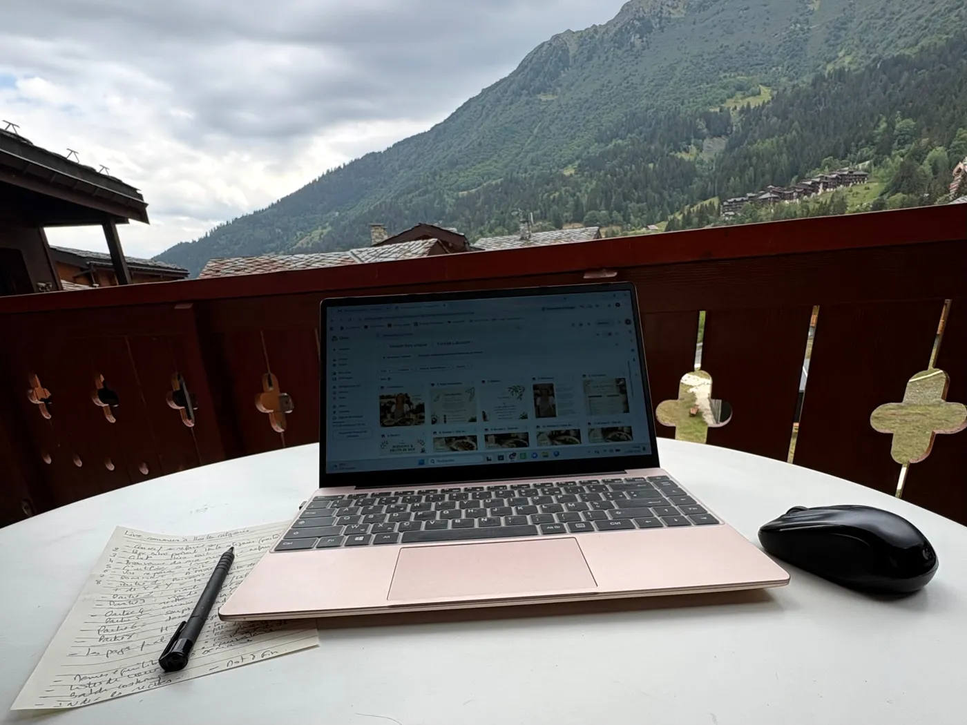 Un ordinateur ouvert sur les pages de Plaisir et Équilibre et une feuille de notes manuscrites, sur une table de balcon face aux montagnes